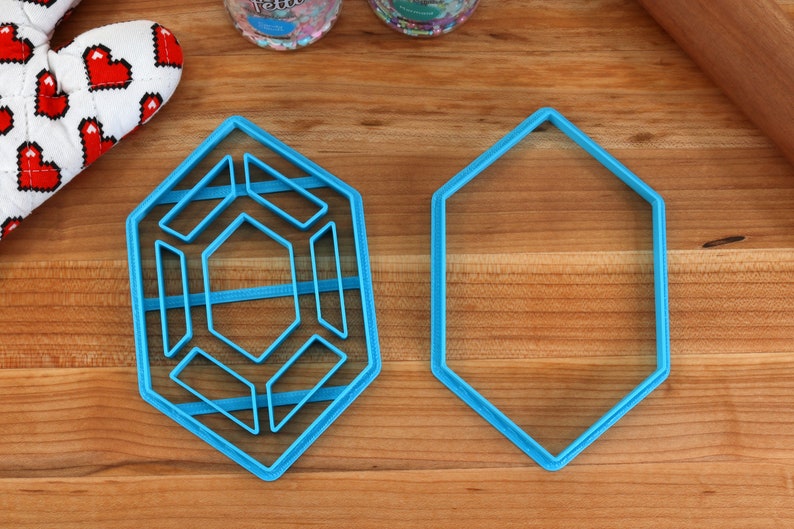 Op de afbeelding: Twee blauwe plastic koekjesvormen op een houten oppervlak. E&eacute;n vorm is een zeshoek, de andere is een complexere geometrische vorm met meerdere lagen.