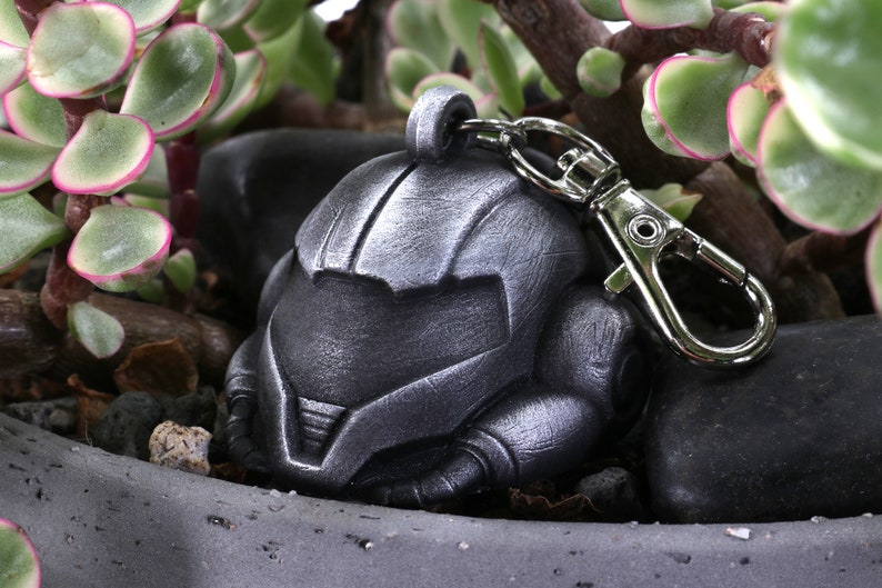 Puede incluir: Un llavero de metal de color plateado con un dise&ntilde;o de casco detallado. El llavero tiene un cierre de metal plateado.