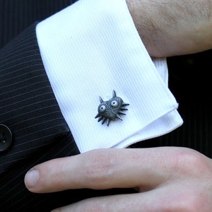 May include: A pair of silver cufflinks featuring a detailed, stylized design of a creature with large eyes and sharp teeth. The cufflinks are worn on a white dress shirt.