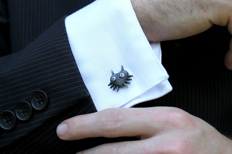May include: A pair of silver cufflinks featuring a detailed, stylized design of a creature with large eyes and sharp teeth. The cufflinks are worn on a white dress shirt.