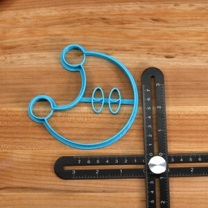 May include: A blue plastic cookie cutter shaped like a crescent moon with two smaller circles inside. The cutter is lying on a wooden surface next to a black ruler.
