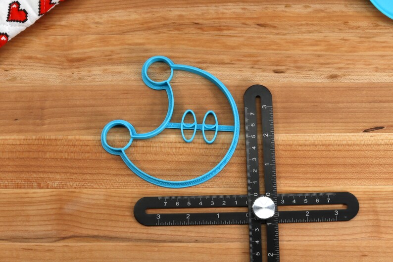 May include: A blue plastic cookie cutter shaped like a crescent moon with two smaller circles inside. The cutter is lying on a wooden surface next to a black ruler.