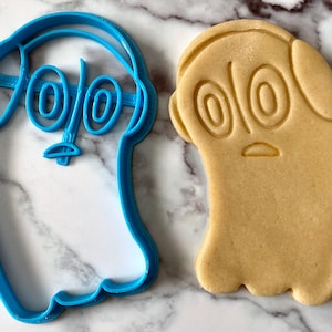 K&ouml;nnte beinhalten: Blauer Plastik-Ausstecher in Form eines Geistes mit gro&szlig;en Augen und Kopfh&ouml;rern. Daneben liegt ein hellbrauner Keks in Form des Ausstechers.