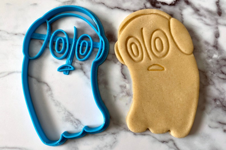 K&ouml;nnte beinhalten: Blauer Plastik-Ausstecher in Form eines Geistes mit gro&szlig;en Augen und Kopfh&ouml;rern. Daneben liegt ein hellbrauner Keks in Form des Ausstechers.