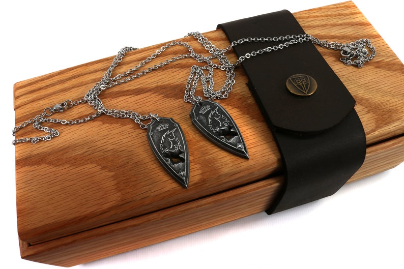 May include: Two silver necklaces with shield-shaped pendants featuring a unicorn design, displayed in a wooden box with a black leather strap and a metal clasp.