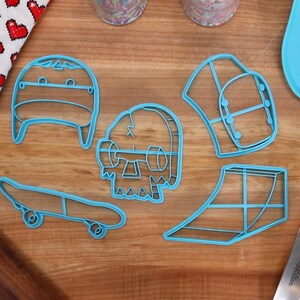 May include: Five blue plastic cookie cutters shaped like a skateboard, a skull, a hat, a ramp, and a helmet. The cutters are on a wooden surface.