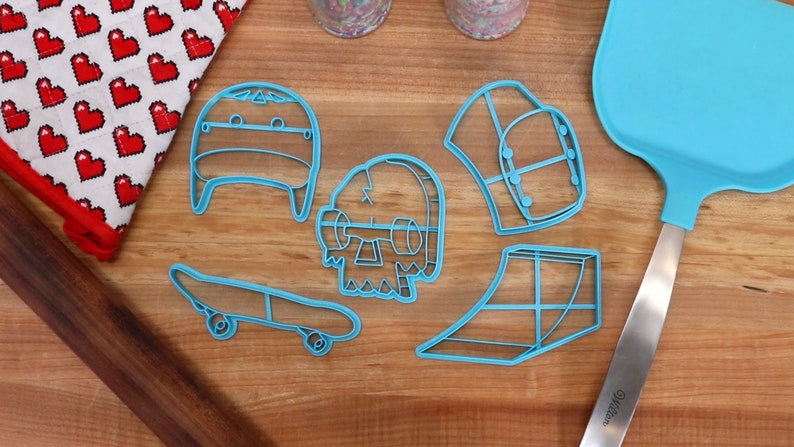 May include: Five blue plastic cookie cutters shaped like a skateboard, a skull, a hat, a ramp, and a helmet. The cutters are on a wooden surface.