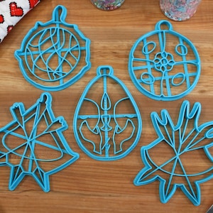 May include: Five blue plastic cookie cutters shaped like a star, a circle, an egg, a snowflake, and a circle with a design inside. The cutters are on a wooden surface.
