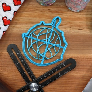 May include: A blue plastic cookie cutter shaped like a ball of yarn. The cutter is on a wooden surface with a ruler and a spatula.