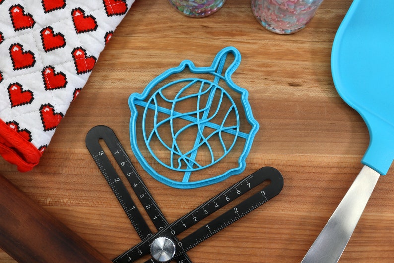 May include: A blue plastic cookie cutter shaped like a ball of yarn. The cutter is on a wooden surface with a ruler and a spatula.