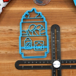 May include: A blue plastic cookie cutter shaped like a gumball machine with a star on top. The gumball machine has a window with gumballs inside and a small star and a bottle with a leaf on the bottom.