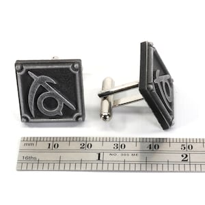 May include: A pair of silver cufflinks with a black and silver design. The design features a stylized symbol with a curved line and a circle.
