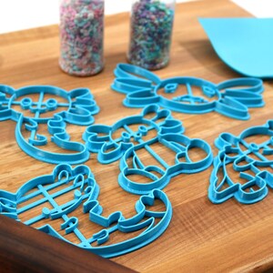 May include: A set of six blue plastic cookie cutters shaped like axolotls. The cutters are on a wooden cutting board with a rolling pin and a spatula.