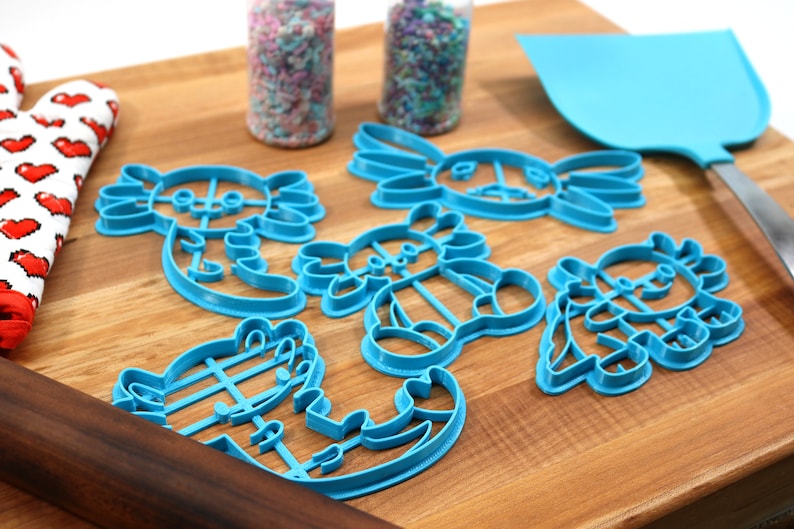 May include: A set of six blue plastic cookie cutters shaped like axolotls. The cutters are on a wooden cutting board with a rolling pin and a spatula.