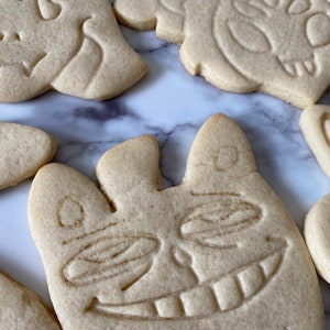 May include: Five sugar cookies decorated with various designs. The cookies are light brown and have a slightly rough texture. One cookie has a smiling face with large eyes and a wide mouth. Another cookie has a simple design with a few lines and curves. The cookies are arranged on a white marble surface.