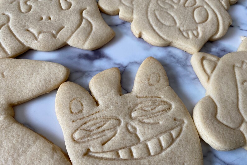 May include: Five sugar cookies decorated with various designs. The cookies are light brown and have a slightly rough texture. One cookie has a smiling face with large eyes and a wide mouth. Another cookie has a simple design with a few lines and curves. The cookies are arranged on a white marble surface.