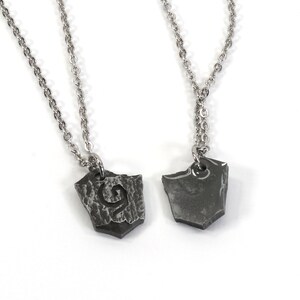 May include: Two silver chain necklaces with matching pendants. The pendants are made of dark metal and have a rough, textured surface. One pendant has a stylized letter 'G' etched into it.