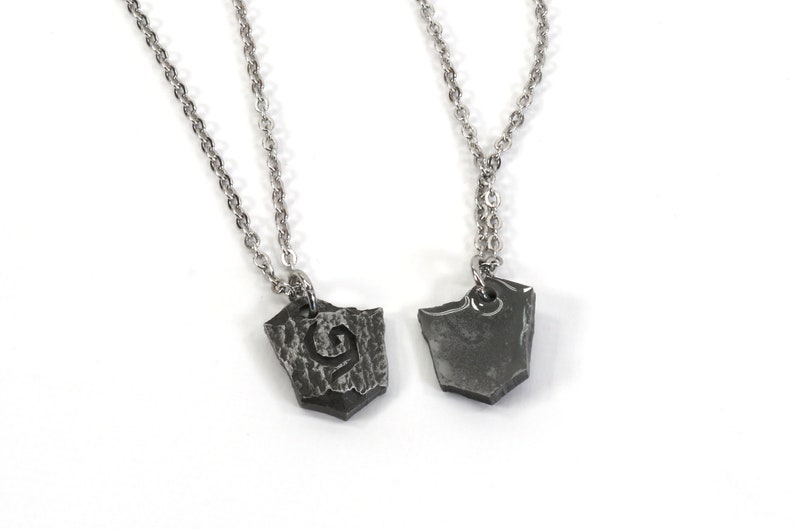 May include: Two silver chain necklaces with matching pendants. The pendants are made of dark metal and have a rough, textured surface. One pendant has a stylized letter 'G' etched into it.
