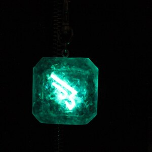 May include: A green, faceted, crystal pendant with a glowing white symbol inside. The pendant is hanging from a chain.