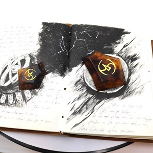 May include: Two brown, geometric, resin bookends with a gold symbol on each. The bookends are on an open notebook with black ink drawings of constellations and other abstract designs.