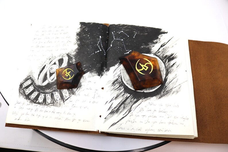 May include: Two brown, geometric, resin bookends with a gold symbol on each. The bookends are on an open notebook with black ink drawings of constellations and other abstract designs.
