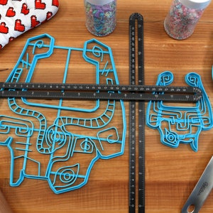 Peut inclure: Deux emportes-pi&egrave;ces en plastique bleu en forme de personnage de jeu vid&eacute;o sur une surface en bois avec une r&egrave;gle, un rouleau &agrave; p&acirc;tisserie et une spatule. Le plus grand emporte-pi&egrave;ce mesure 30,5 cm de large et le plus petit 15,2 cm de large.