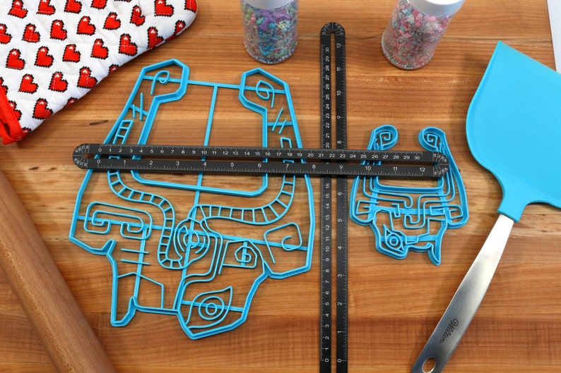 Peut inclure: Deux emportes-pi&egrave;ces en plastique bleu en forme de personnage de jeu vid&eacute;o sur une surface en bois avec une r&egrave;gle, un rouleau &agrave; p&acirc;tisserie et une spatule. Le plus grand emporte-pi&egrave;ce mesure 30,5 cm de large et le plus petit 15,2 cm de large.