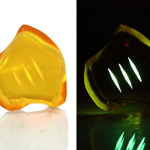 May include: A translucent, amber-colored, abstract sculpture with three diagonal, etched lines. The sculpture glows with a bright green light in a dark setting, creating a striking visual contrast.