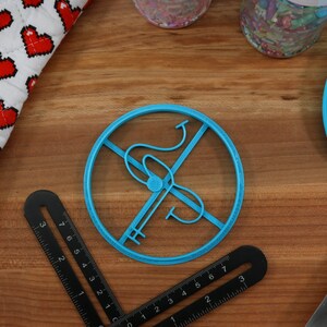 May include: A blue plastic cookie cutter shaped like a circle with a stylized letter "S" inside. The cutter is on a wooden surface.