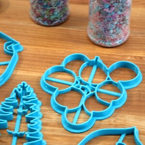 May include: A set of blue plastic cookie cutters in various shapes, including a Christmas tree, a snowflake, a bird, and a leaf. The cutters are on a wooden surface.