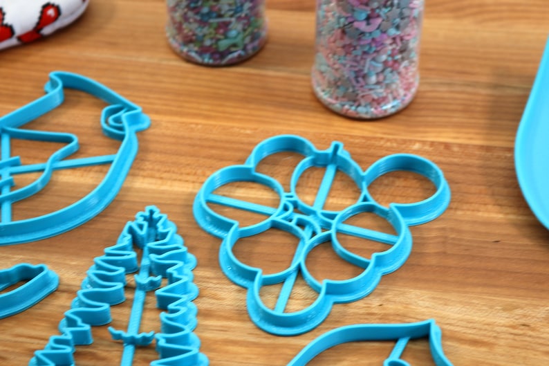 May include: A set of blue plastic cookie cutters in various shapes, including a Christmas tree, a snowflake, a bird, and a leaf. The cutters are on a wooden surface.