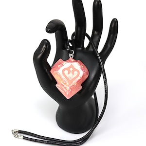 May include: A pink and white pendant necklace with a geometric design. The pendant is shaped like a diamond and has a white design in the centre. The necklace is made of black cord.