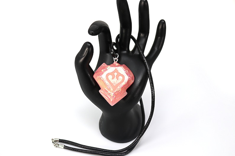 May include: A pink and white pendant necklace with a geometric design. The pendant is shaped like a diamond and has a white design in the centre. The necklace is made of black cord.