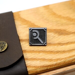 Peut inclure: Une &eacute;pingle en m&eacute;tal argent&eacute; avec un motif carr&eacute; noir repr&eacute;sentant un symbole stylis&eacute;. L'&eacute;pingle est sur une surface en bois.