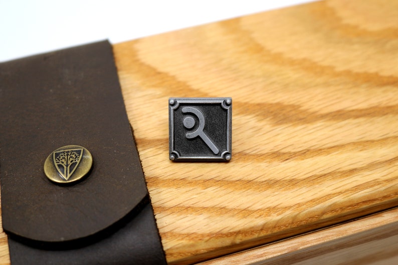 Peut inclure: Une &eacute;pingle en m&eacute;tal argent&eacute; avec un motif carr&eacute; noir repr&eacute;sentant un symbole stylis&eacute;. L'&eacute;pingle est sur une surface en bois.