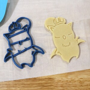 May include: Two cookie cutters, one blue and one beige, shaped like a cartoon character wearing a crown. The cutters are on a white surface.