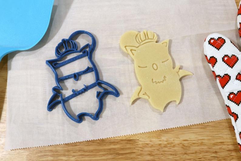 May include: Two cookie cutters, one blue and one beige, shaped like a cartoon character wearing a crown. The cutters are on a white surface.