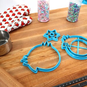 May include: A set of blue plastic cookie cutters in the shapes of a star, a circle with a cross inside, and a round shape with a wavy edge. The cutters are on a wooden cutting board with a ruler, a rolling pin, a measuring cup, and two jars of sprinkles.
