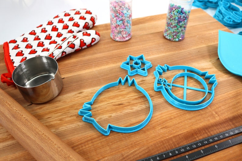 May include: A set of blue plastic cookie cutters in the shapes of a star, a circle with a cross inside, and a round shape with a wavy edge. The cutters are on a wooden cutting board with a ruler, a rolling pin, a measuring cup, and two jars of sprinkles.
