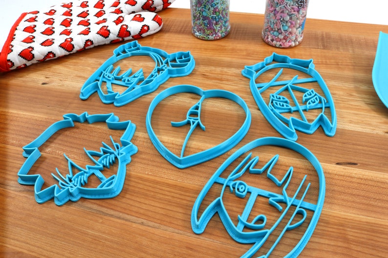 May include: Five blue plastic cookie cutters shaped like anime characters and a heart. The cutters are on a wooden surface.