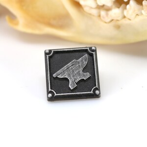 May include: A small, square, black metal pin with a silver anvil design. The pin has a textured surface and a raised border.