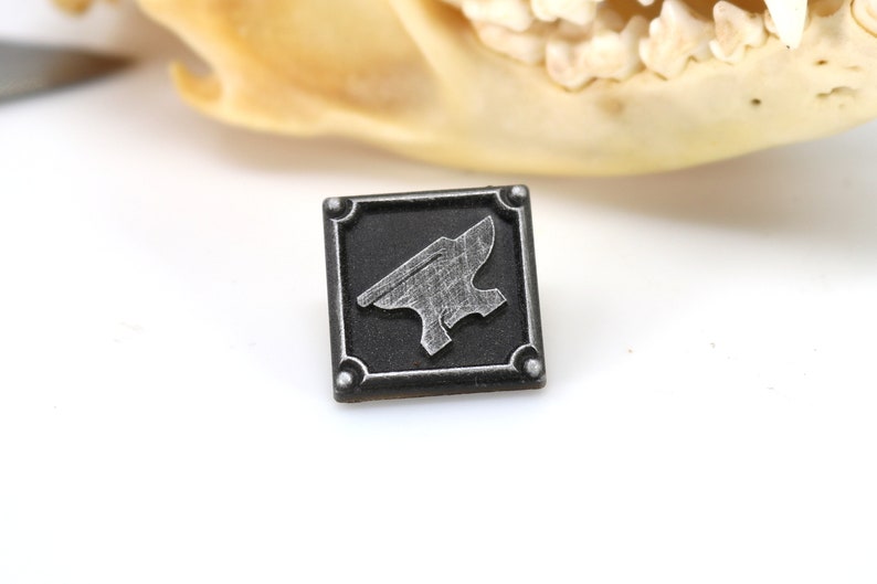 May include: A small, square, black metal pin with a silver anvil design. The pin has a textured surface and a raised border.