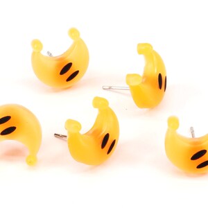 以下が含まれることがあります： 黒い目をつけた黄色の三日月型のイヤリングが5つセットになっています。イヤリングはプラスチック製で、銀色のポストが付いています。