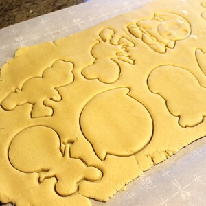 May include: A sheet of cookie dough with various shapes cut out, including a ghost, a speech bubble, and a few other unidentified shapes. The dough is on a white parchment paper with a grid pattern.
