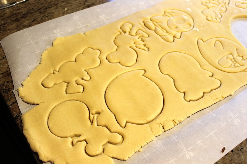 May include: A sheet of cookie dough with various shapes cut out, including a ghost, a speech bubble, and a few other unidentified shapes. The dough is on a white parchment paper with a grid pattern.