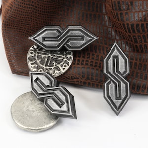 May include: Four silver-colored metal charms in the shape of the "S" symbol, with a textured finish. The charms are displayed with a brown leather pouch and a silver coin. The background is white.