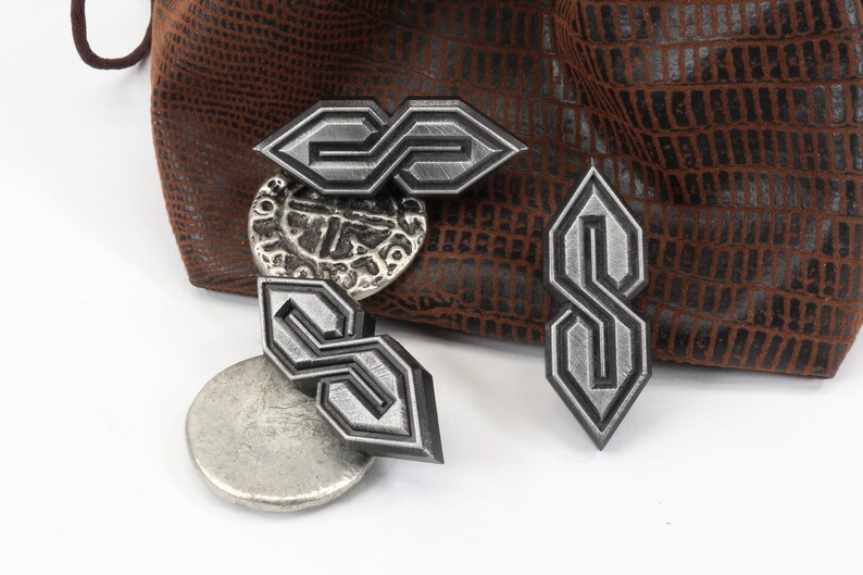 May include: Four silver-colored metal charms in the shape of the "S" symbol, with a textured finish. The charms are displayed with a brown leather pouch and a silver coin. The background is white.