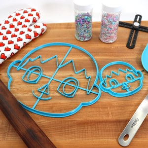 May include: A wooden cutting board with a blue cookie cutter in the shape of a face, a smaller blue cookie cutter in the shape of a face, a rolling pin, a blue spatula, two jars of sprinkles, and a red and white oven mitt.