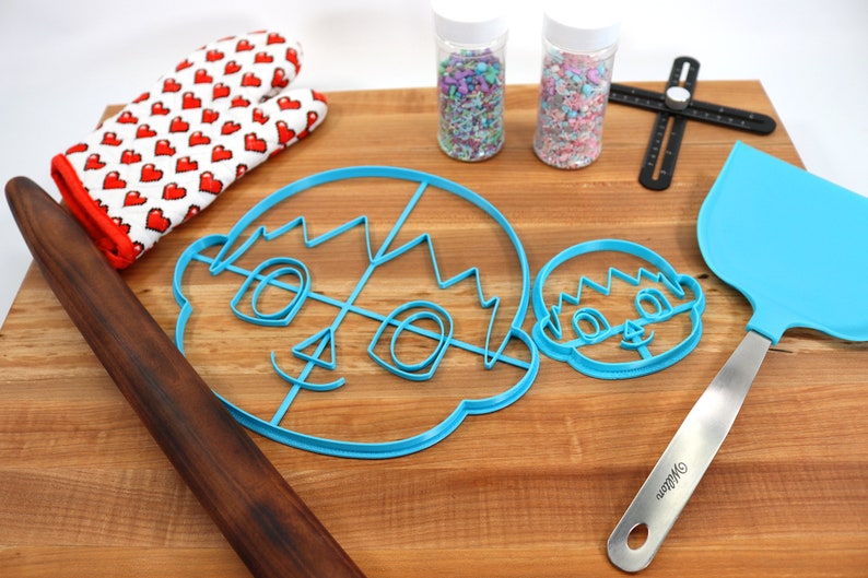 May include: A wooden cutting board with a blue cookie cutter in the shape of a face, a smaller blue cookie cutter in the shape of a face, a rolling pin, a blue spatula, two jars of sprinkles, and a red and white oven mitt.