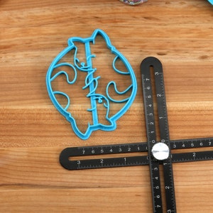 May include: A blue plastic cookie cutter shaped like two cats facing each other. The cutter is on a wooden surface with a black ruler.